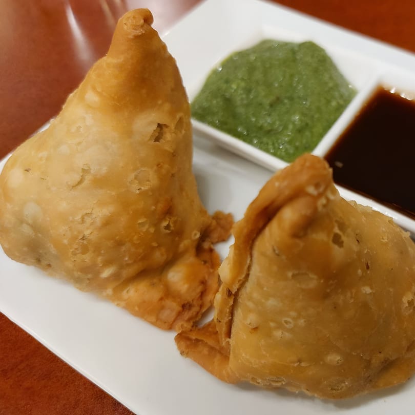 Two golden-brown samosas with green and dark chutneys on a white plate at Roti Indian Kitchen.